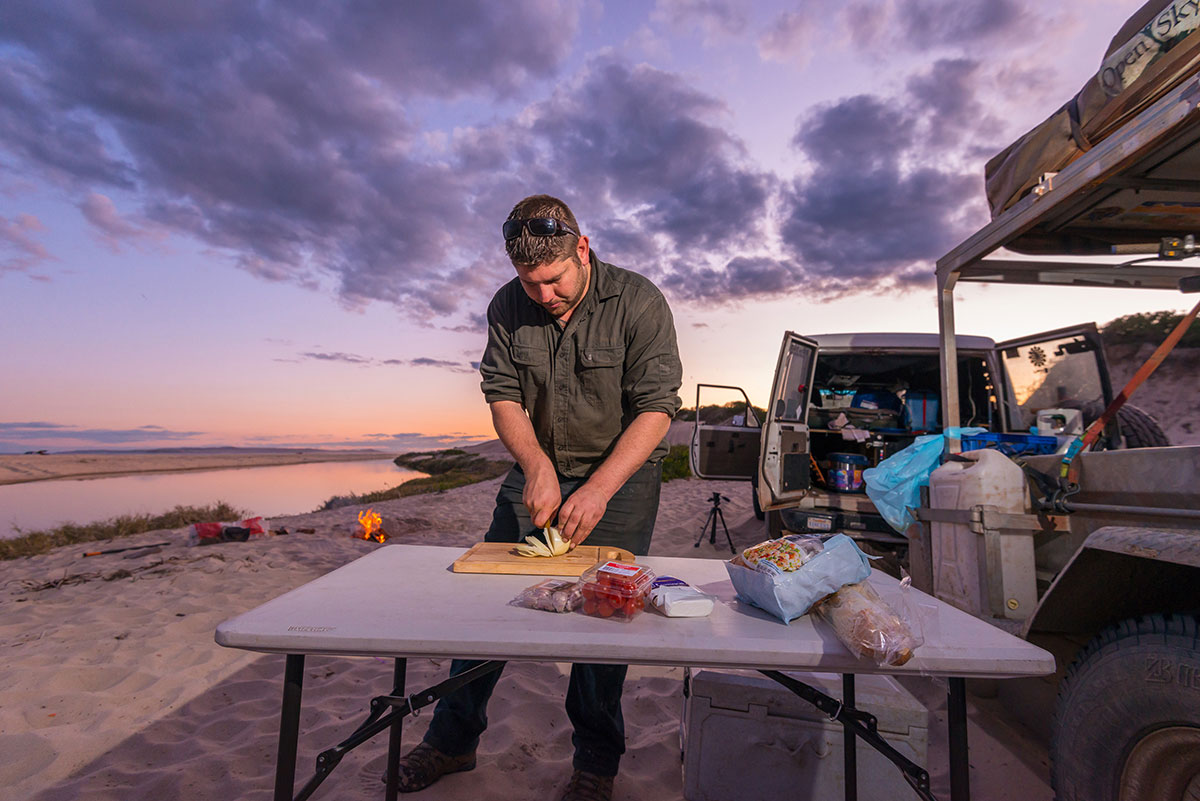 Man-preparing-food-next-to-a-camper-trailer-at-the-beach